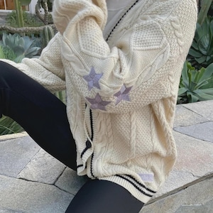 May include: Cream-colored cable knit cardigan with black and white striped trim. The sweater features three lavender star patches on the left sleeve. The cardigan has a button-up front and a hood.