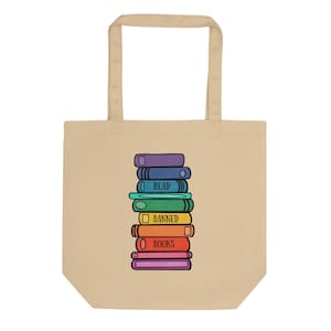 May include: A beige tote bag featuring a centered graphic of a vertical stack of colorful books. The book spines are purple, blue, teal, green, yellow, orange, red, and pink. The text READ BANNED BOOKS is printed in black on the spines.