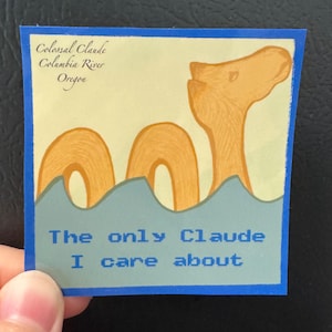 May include: A square sticker with a blue border featuring a tan illustration of a sea creature emerging from blue water. Text reads: Colossal Claude Columbia River Oregon and The only Claude I care about in blue and black lettering.