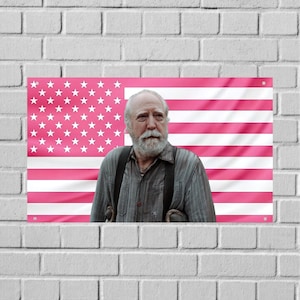 May include: A rectangular wall hanging featuring a portrait of a man with a white beard and suspenders, set against a pink American flag background. The flag has white stars and pink stripes. The wall hanging is mounted on a white brick wall.