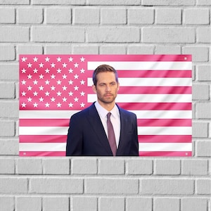 May include: A rectangular wall hanging featuring a pink American flag with white stars and stripes. A man in a navy suit, white shirt, and burgundy tie stands in front of the flag. The wall hanging is mounted on a white brick wall.