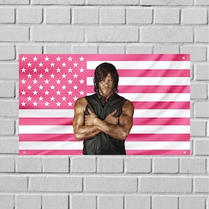 Puede incluir: Una bandera estadounidense rosa y blanca con un retrato de un hombre con los brazos cruzados. El hombre lleva un chaleco negro y tiene suciedad en los brazos y la cara. La bandera está colgada en una pared de ladrillos blancos.