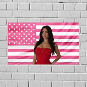 Puede incluir: Una persona con un vestido rojo sin tirantes con un detalle de ojo de cerradura frente a una bandera estadounidense rosa. La bandera tiene estrellas blancas y rayas rosas. La imagen está en una pared de ladrillos blancos.