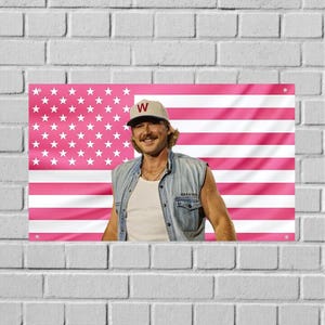 May include: A rectangular wall flag featuring a pink and white American flag pattern with a central photographic print of a person wearing a beige baseball cap with a red W, a white tank top, and a light blue denim vest over a white shirt.