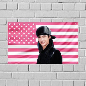 Op de afbeelding: Een roze en witte Amerikaanse vlag met een portret van een persoon met een zwarte hoed en jas. De vlag is bevestigd aan een witte bakstenen muur.