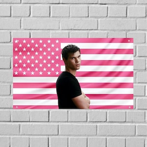 Puede incluir: Un estandarte de pared rectangular que presenta un diseño de bandera estadounidense estilizado con rayas horizontales alternas blancas y rosas, y un campo azul con estrellas blancas. Una persona de pelo corto oscuro viste una camiseta negra.