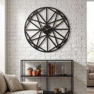 May include: A large, circular wall clock featuring a black metal frame with a geometric, spoke-like pattern. The clock has a solid black center and thin, metallic gold-toned hour and minute hands, creating a stark contrast against the dark frame.