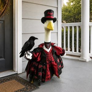 May include: A white goose figurine dressed as a vampire, wearing a black top hat with a red bat emblem, a black and red cape, and a bat-patterned skirt. A black raven figurine is perched on one of the goose's white wings, and the goose wears a collar.