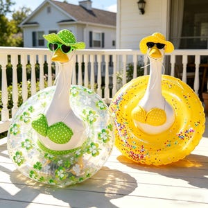 May include: Two inflatable goose pool floats, one with a green polka dot hat, sunglasses, and bikini, and the other with a yellow hat, sunglasses, and bikini. The floats are on a wooden deck with a white railing and a house in the background.