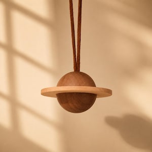 May include: A hanging ornament shaped like a planet with a dark brown wooden sphere and a lighter beige wooden ring encircling the center. A thin brown leather cord is attached to the top of the sphere for suspension, creating a simple geometric design.