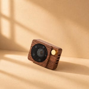 May include: A rectangular portable speaker with a dark brown wood grain finish, featuring a circular black mesh speaker grille on the left side and a single polished gold-colored circular control knob on the right side of the front panel.