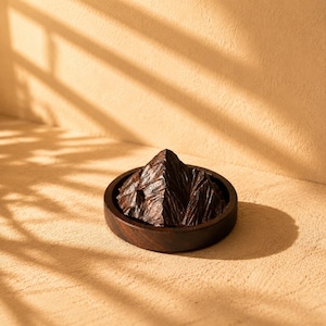 May include: A dark brown wooden sculpture shaped like a jagged mountain peak, resting inside a circular dark brown wooden tray. The sculpture features sharp, angular facets and a deep, rich wood grain texture, creating a mountain-like form.
