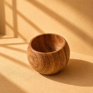 May include: A small, round wooden bowl with a natural light brown finish and visible wood grain patterns. The bowl features a smooth, curved exterior and a deep, open interior, showcasing the organic texture and warm tones of the wood material.