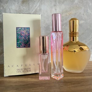 May include: An assortment of perfume bottles and a box. The box is cream-colored with the word "ACAPELLA" and a colorful floral design. The bottles are various shapes and sizes, with pink and gold accents. One bottle is labeled "EAU DE TOILETTE 1.9 FL. OZ. / 56 mL."