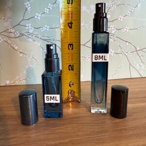 Pode incluir: Dois frascos de perfume de vidro azul, um rotulado "5ML" e outro "8ML", com borrifadores pretos e tampas correspondentes. Uma fita m&eacute;trica amarela &eacute; colocada entre os frascos.