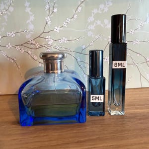 Pode incluir: Tr&ecirc;s frascos de perfume azuis de tamanhos variados. O frasco maior tem uma tampa prateada. Os frascos menores est&atilde;o rotulados como "5ML" e "8ML". Os frascos est&atilde;o sobre uma superf&iacute;cie de madeira com um fundo floral.