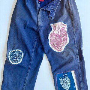 May include: Blue work pants with multiple patches. Patches include a pink heart design, a blue and white abstract design, and a blue and white design. The pants have a button fly and a belt loop.