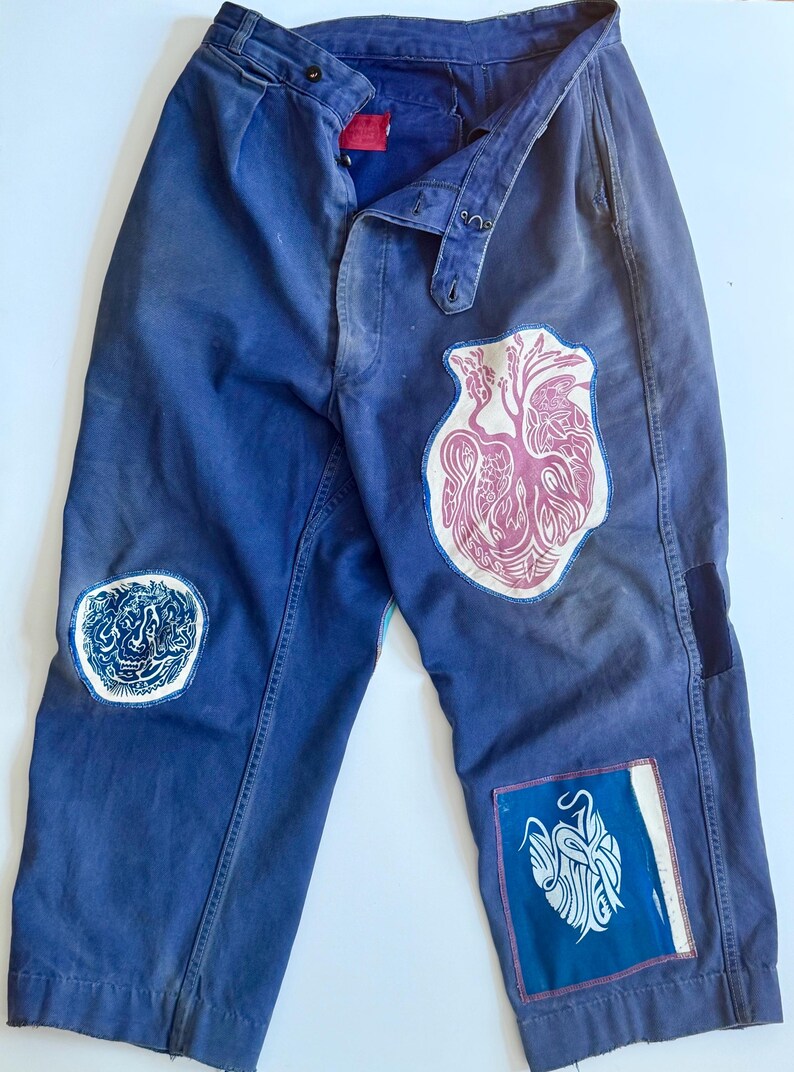 May include: Blue work pants with multiple patches. Patches include a pink heart design, a blue and white abstract design, and a blue and white design. The pants have a button fly and a belt loop.