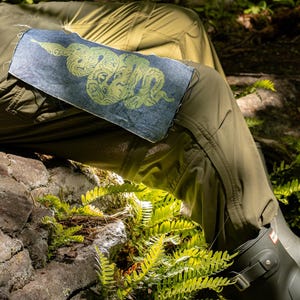 May include: A denim patch featuring a green snake design, sewn onto olive green pants. The snake is intricately detailed with a swirling pattern. The image is set outdoors with ferns and a rock in the background.