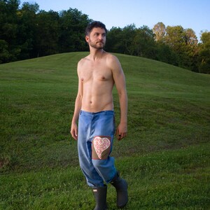 May include: Blue denim pants with a heart-shaped patch on the knee. The patch is a light brown color with a white design. The person is wearing black rubber boots and standing in a grassy field.