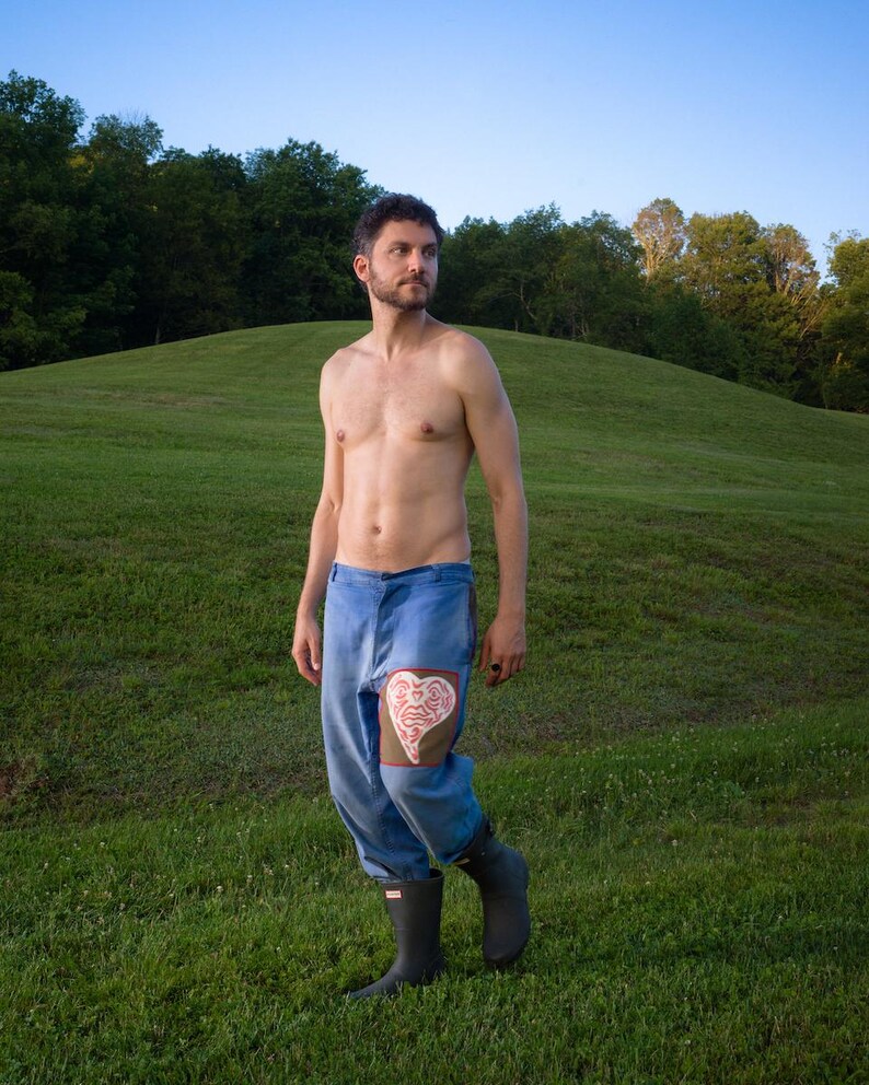May include: Blue denim pants with a heart-shaped patch on the knee. The patch is a light brown color with a white design. The person is wearing black rubber boots and standing in a grassy field.