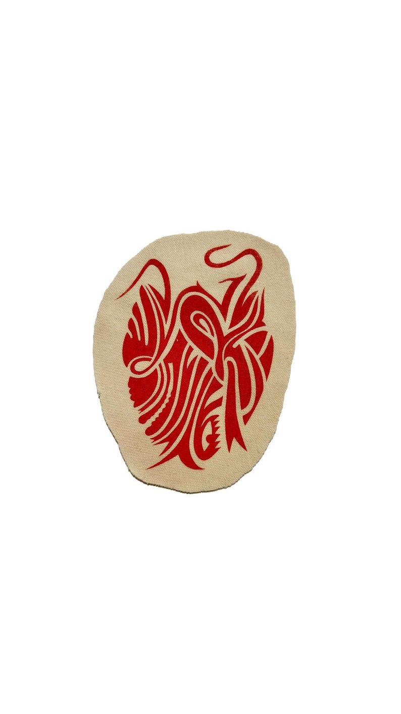 May include: A beige, oval-shaped patch featuring a vibrant red tribal design. The design is intricate, with flowing lines and stylized shapes. The patch is likely for decorative purposes, possibly for clothing or accessories.