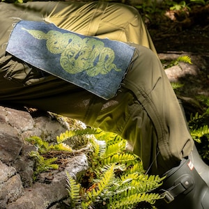 May include: A denim patch with a green snake design is sewn onto olive green pants. The snake is stylized with intricate patterns. The patch is rectangular with frayed edges. The image is set outdoors with ferns and a rock in the background.