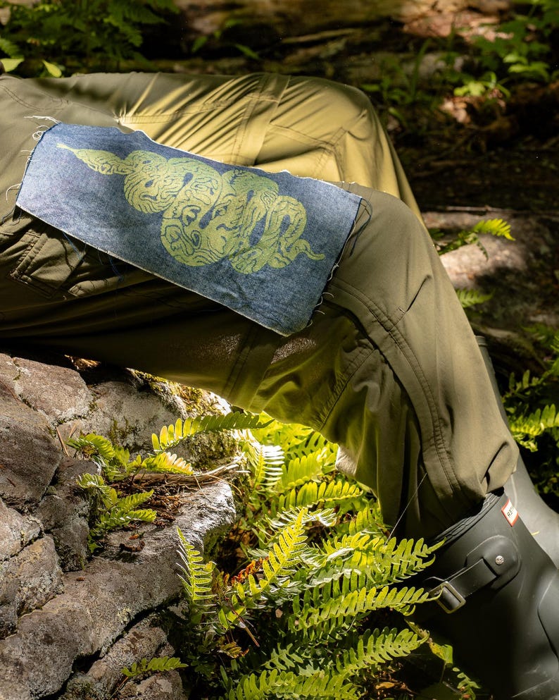 May include: A denim patch with a green snake design is sewn onto olive green pants. The snake is stylized with intricate patterns. The patch is rectangular with frayed edges. The image is set outdoors with ferns and a rock in the background.