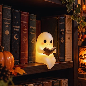 May include: A small, glowing, white ghost-shaped lamp with a smooth, rounded form, black eyes, and pink cheeks, sitting on a shelf while holding a small, open black book. The lamp emits a warm, soft light, illuminating the surrounding vintage books.