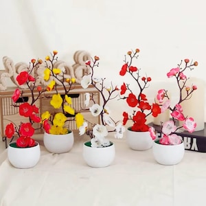 May include: Arrangement of artificial flowering branches in small white ceramic pots. The flowers are in shades of red, yellow, white, and pink. The pots have a green interior and are arranged on a white surface.