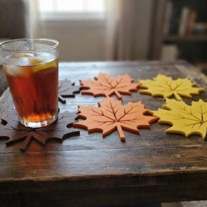 Könnte beinhalten: Ein Set aus fünf ahornblattförmigen Untersetzern in den Farben Dunkelbraun, Orange und Gelb. Ein Untersetzer befindet sich unter einem klaren Glas, das mit Eistee und einer Zitronenscheibe gefüllt ist. Die Untersetzer haben ausgeschnittene Blattadern.
