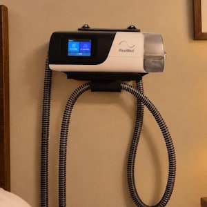 May include: A white and black ResMed CPAP machine mounted on a wall. The device features a digital display screen, a clear water chamber on the right side, and two long, flexible, ribbed gray hoses hanging downward from the base of the unit.