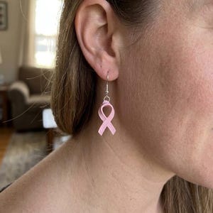 May include: A light pink awareness ribbon earring featuring a smooth, matte finish. The ribbon is suspended from a simple silver-toned metal hook wire, creating a dangling design that hangs vertically from the earlobe with a clean, minimalist look.