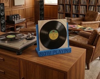 Puede incluir: Un soporte azul para discos de vinilo con el texto NOW PLAYING en relieve en la parte frontal. El soporte sostiene un disco de vinilo negro en una funda de papel blanca, colocado junto a un tocadiscos plateado sobre una superficie de madera.