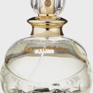 Könnte beinhalten: Eine transparente Parfümflasche aus Glas mit einem goldfarbenen Deckel und einem transparenten, kugelförmigen Verschluss. Die Flasche hat eine abgerundete Form und den Text "NAJ OLEAR" in orangefarbenen Buchstaben. Die Parfümflasche steht vor weißem Hintergrund.