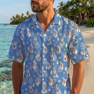 May include: A light blue short-sleeve button-down shirt featuring an all-over print of Olaf the snowman from Frozen, interspersed with white and pink stylized trees and snowflakes. The shirt has a classic collar and a front button closure.