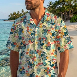May include: A short-sleeved button-down shirt featuring a cream background with a repeating pattern of colourful roosters, green tropical palm leaves, and orange hibiscus flowers. The text "HEI HEI" is printed in red throughout the fabric design.