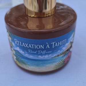 Peut inclure: Un flacon diffuseur à roseaux cylindrique marron avec un bouchon doré. L'étiquette présente une scène de plage tropicale avec de l'eau bleue, des feuilles de palmier et des fleurs, avec le texte "RELAXATION À TAHITI" et "Reed Diffuser".