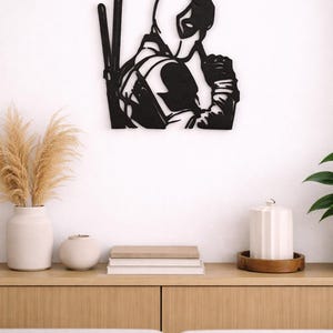 Può includere: Un'opera d'arte da parete in metallo nero che presenta la silhouette stilizzata di un personaggio mascherato con due spade legate sulla schiena. Il design mostra il personaggio di profilo con una mano vicino al viso, in uno stile piatto.