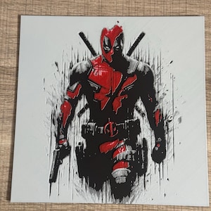 Può includere: Una stampa quadrata grigia con un'illustrazione stilizzata di Deadpool. Il personaggio è raffigurato in nero e rosso, con una pistola e due katane. Lo sfondo ha un effetto strutturato e macchiato.