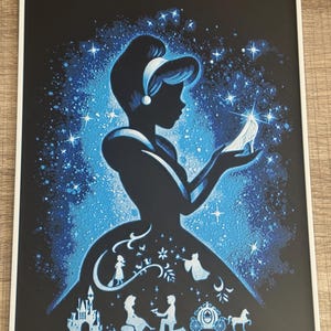 Op de afbeelding: Een ingelijste kunstdruk met een silhouet van Assepoester die een glazen muiltje vasthoudt. Het zwarte silhouet contrasteert met een blauw-witte sterrenachtergrond. Het jurkontwerp bevat silhouetten van een kasteel, koets en andere sprookjeselementen.