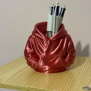 Peut inclure: Un porte-stylo rouge métallique en forme de sweat à capuche avec une poche avant. Le porte-stylo est rempli de plusieurs stylos blancs avec des pointes d'encre colorées. Le porte-stylo est posé sur une surface en bois clair.