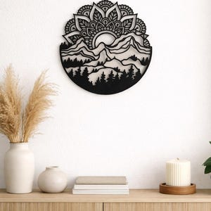 Peut inclure: Décoration murale en métal noir représentant un paysage de montagnes et de forêts avec un soleil et un motif floral de mandala. L'œuvre d'art circulaire est fixée sur un mur blanc au-dessus d'un meuble en bois. Autres éléments : vase avec plantes séchées, petit vase, livres et bougie.