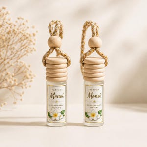 May include: Two glass car air fresheners with wooden tops and rope hangers. Each bottle has a label with the word "Monoï" and a floral illustration. The air fresheners are designed to hang in a vehicle.