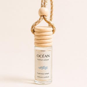 May include: A car air freshener in a clear glass bottle with a wooden top and a woven rope hanger. The label reads "OCÉAN" with "Parfum voiture" and "Fraîcheur océan" below a wave graphic. The wooden parts are light beige.