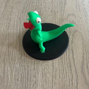Può includere: Statuina di lucertola in argilla verde fatta a mano con lingua rossa, occhi bianchi e piccole zampe. La lucertola è posizionata su una base circolare nera, creando un'estetica stravagante e giocosa. Un oggetto decorativo unico.