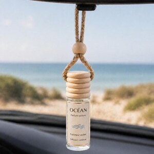 May include: A car air freshener suspended from a rearview mirror. The clear glass bottle has a wooden cap and is labelled "OCÉAN Parfum voiture". The text "Fraîcheur océan" and "Diffusion continue" are also visible. The background shows a beach and the sea.