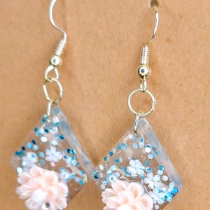 Puede incluir: Un par de pendientes en forma de diamante hechos de resina transparente con purpurina azul y pequeños detalles florales blancos. Cada pendiente tiene una flor tridimensional de color rosa pálido en el centro y cuelga de un gancho de metal plateado.