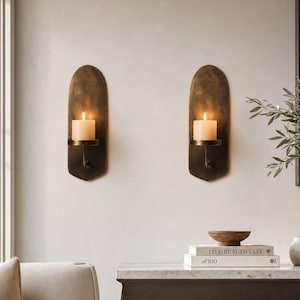 Puede incluir: Dos apliques de pared arqueados con un acabado de bronce oscuro desgastado, cada uno con un pequeño estante circular que sostiene una vela de pilar blanca encendida. Los apliques tienen una parte superior redondeada y una base plana.
