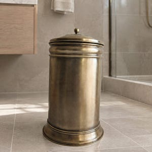 May include: A cylindrical waste bin crafted from antique brass with a brushed metallic finish. The container features a rounded base, a smooth body with subtle horizontal ridges, and a removable lid topped with a small, centered spherical knob.